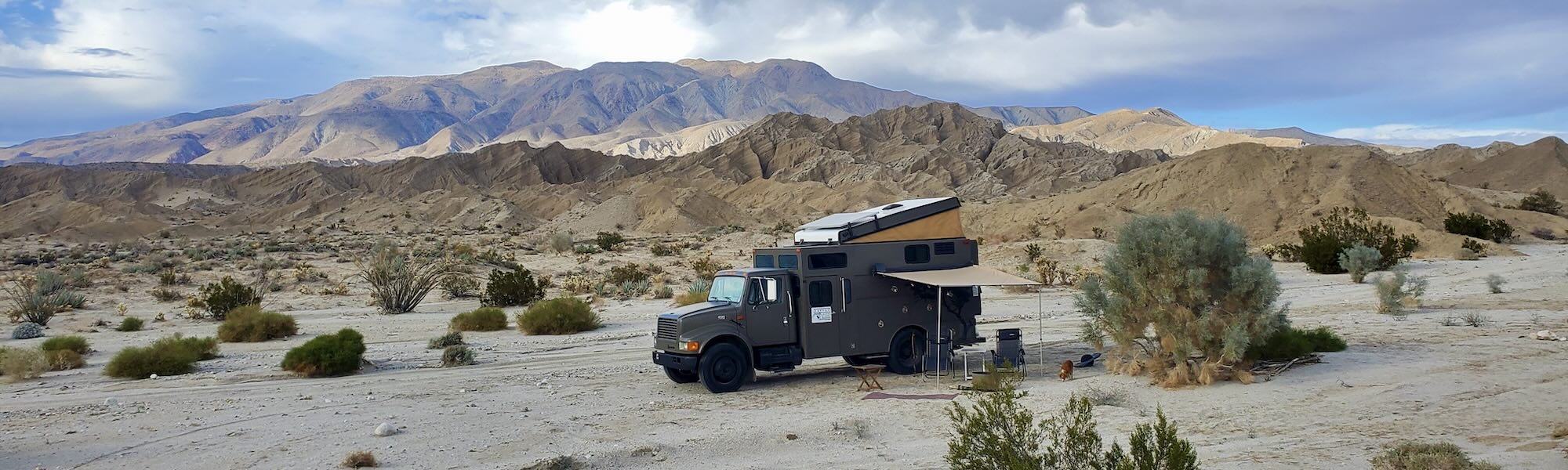 Large olive green truck with popup roof against backdrop of desert with sand and low shrubs