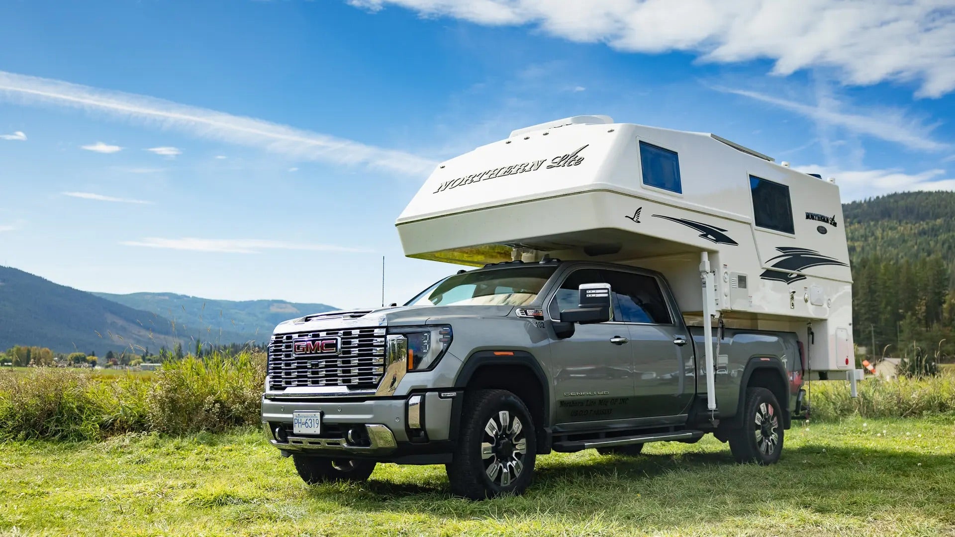Gray pickup truck with a white northern lite camper on a grassy field with mountains in the background