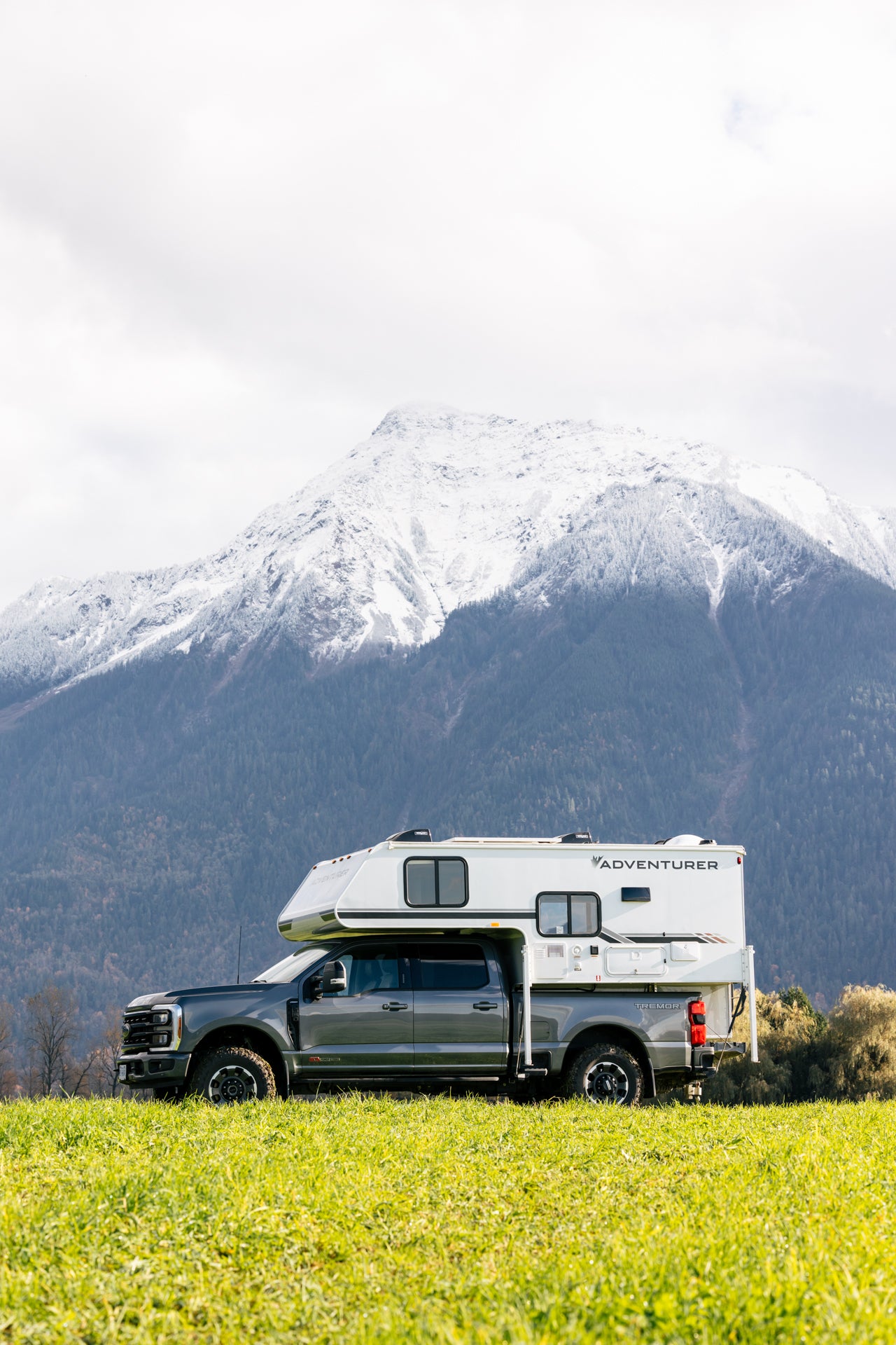 Grey truck with white Adventurer 80RB camper parked in a field with snow-capped mountains in the background
