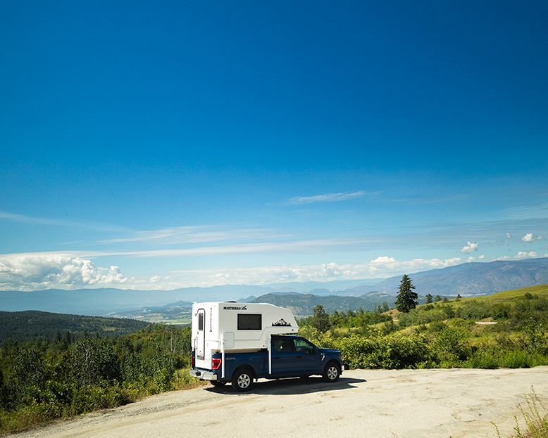 Navy truck with a white Northern Lite 610 camper on a scenic road with mountains in the background