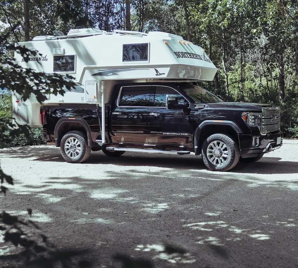 Black truck with a white Sportsman Plus Northern Lite camper parked on a road surrounded by trees