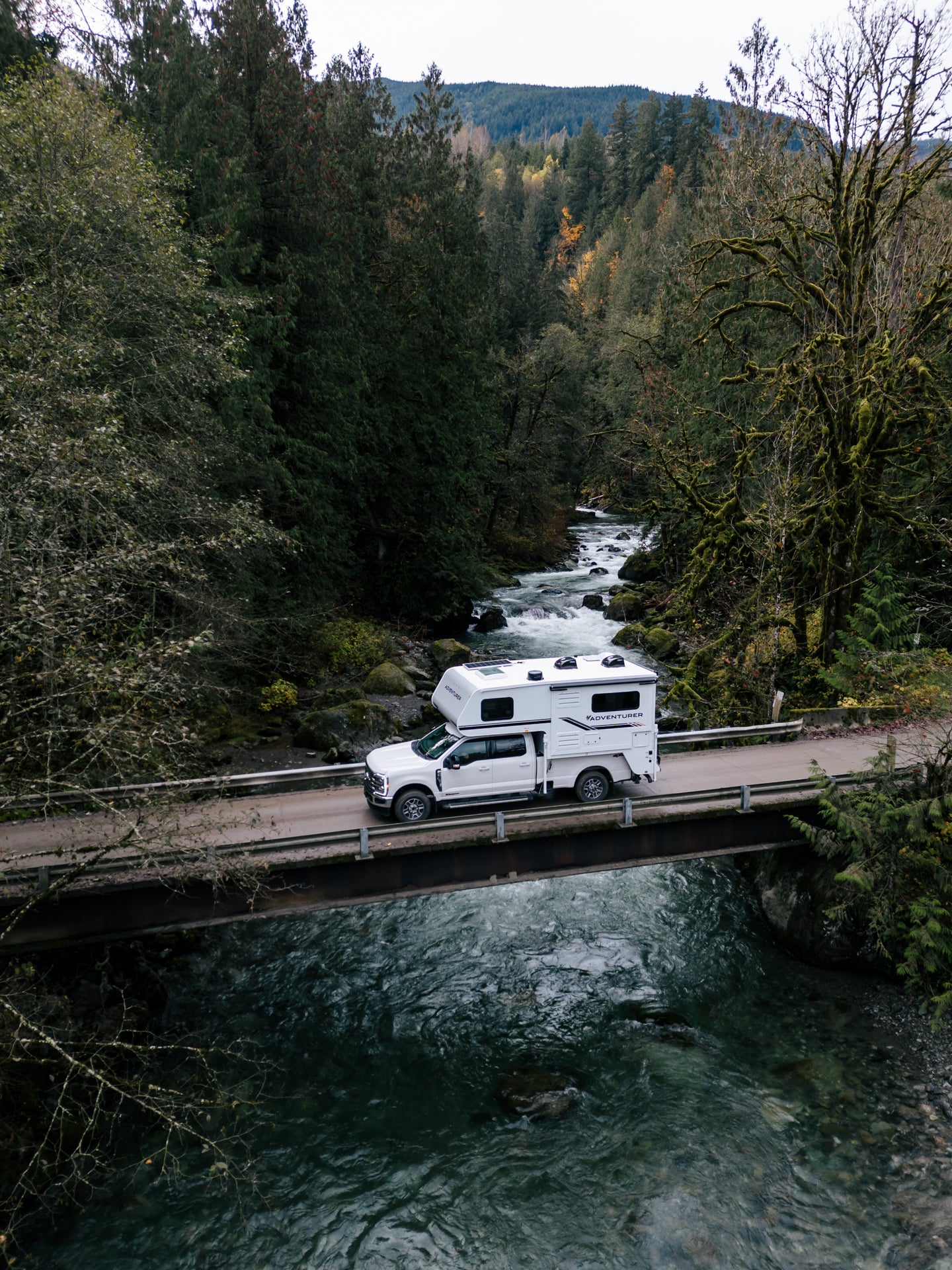 White truck with a white 89RBS Adventurer truck camper on a scenic road surrounded by trees and a river