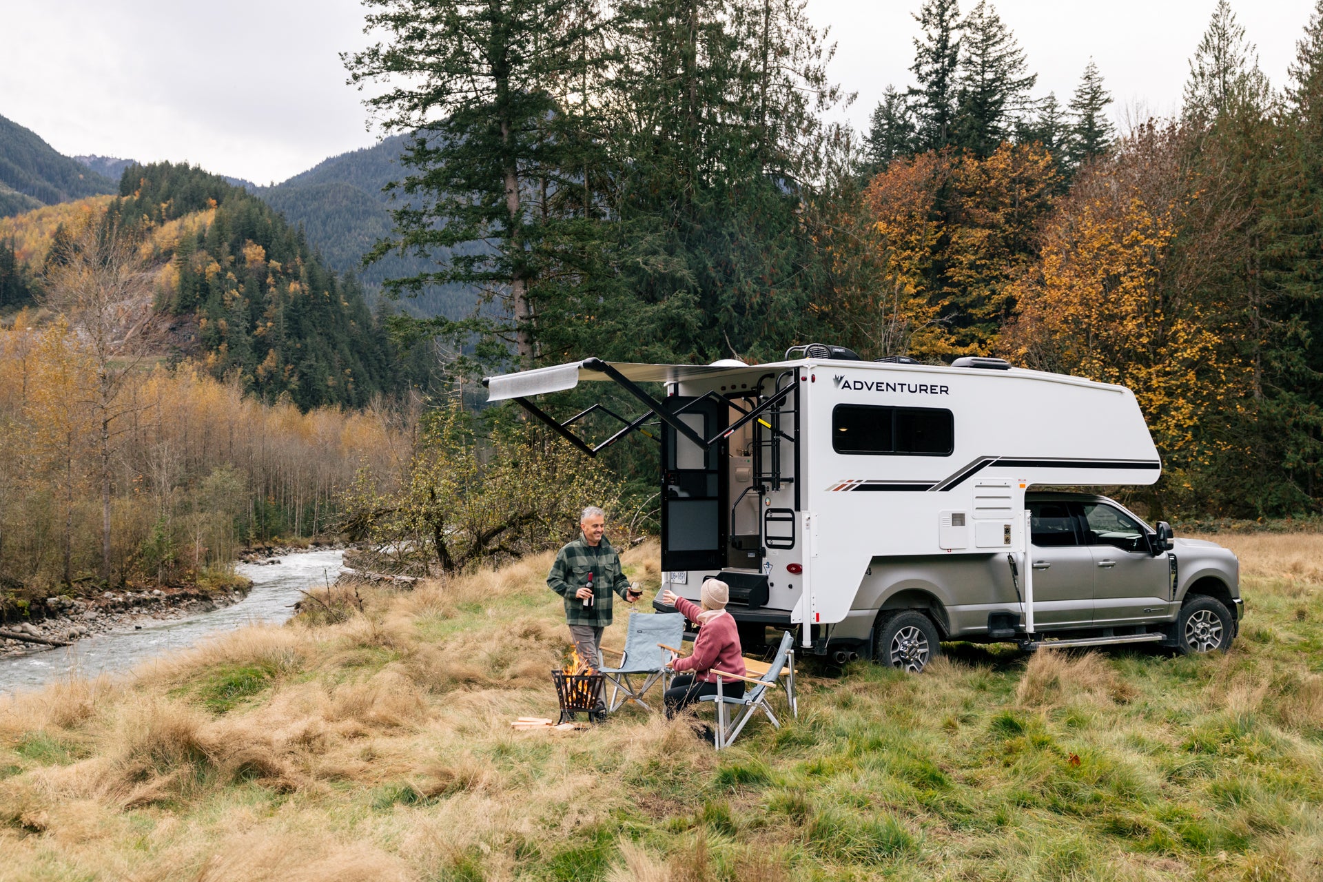 Two people with Dometic chairs by a white 89RB Adventurer truck camper in a forested area with mountains in the background