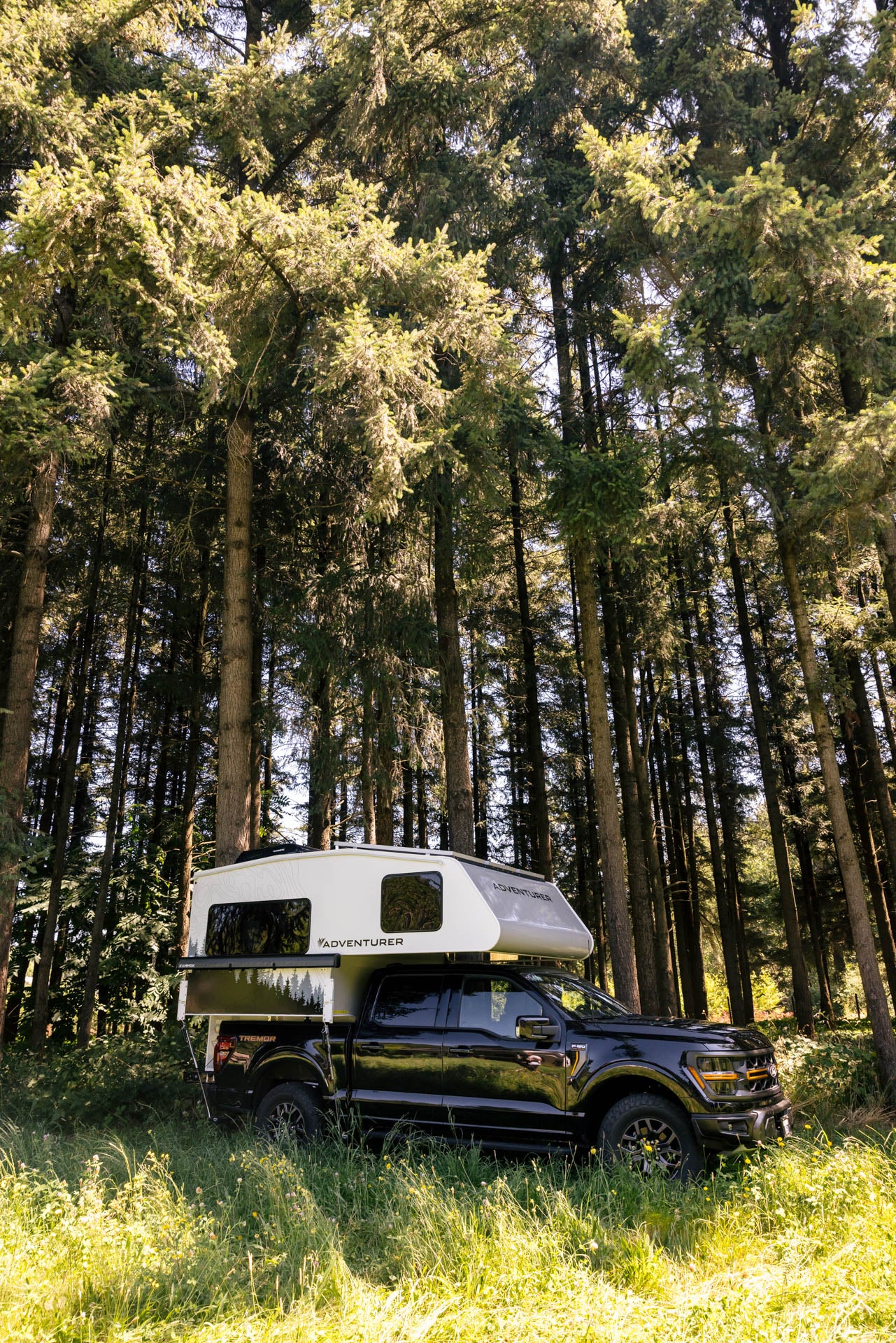 Black crew cab truck with a white Adventurer 65RB camper shell and Kammok awning parked in a forest