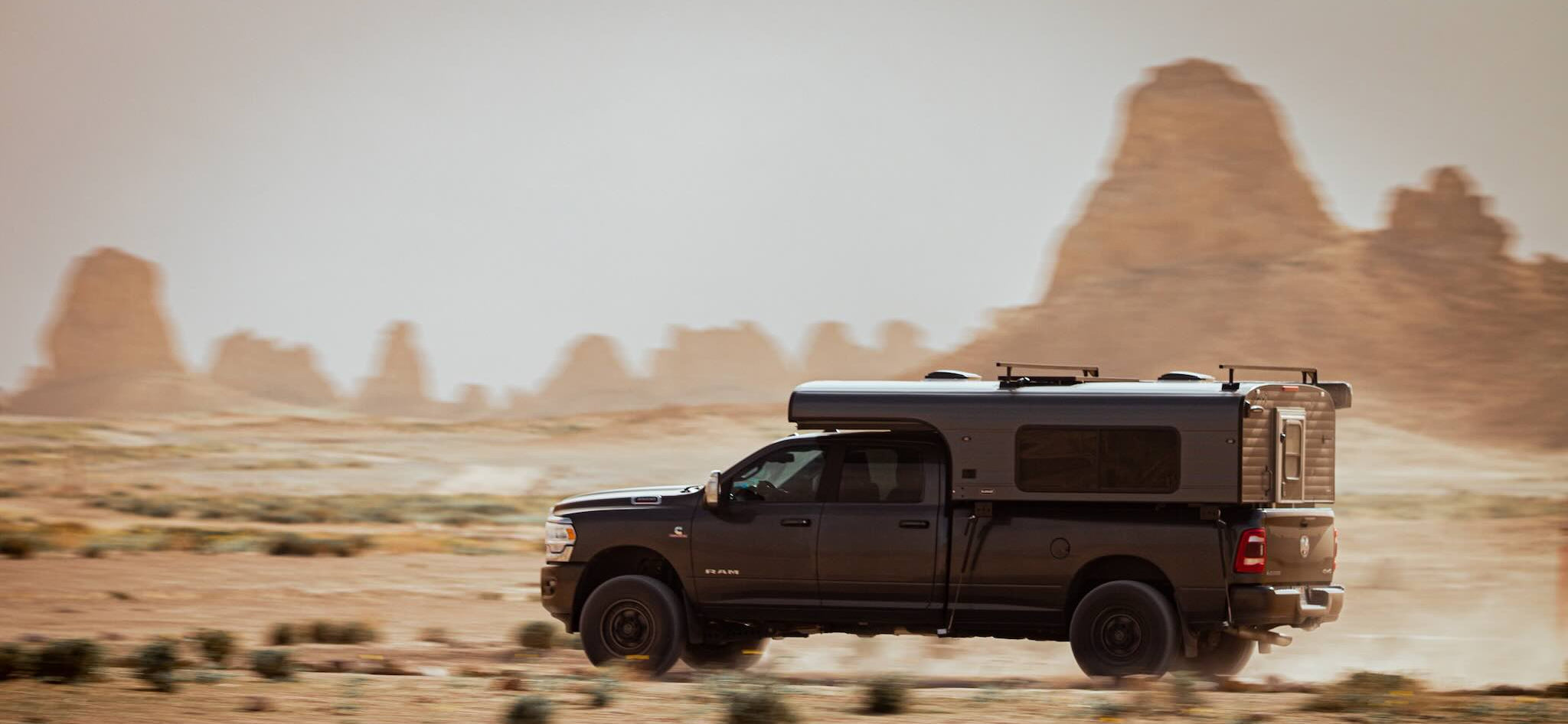 Black truck with grey Alaskan cabover camper driving through a desert landscape with rock formations.