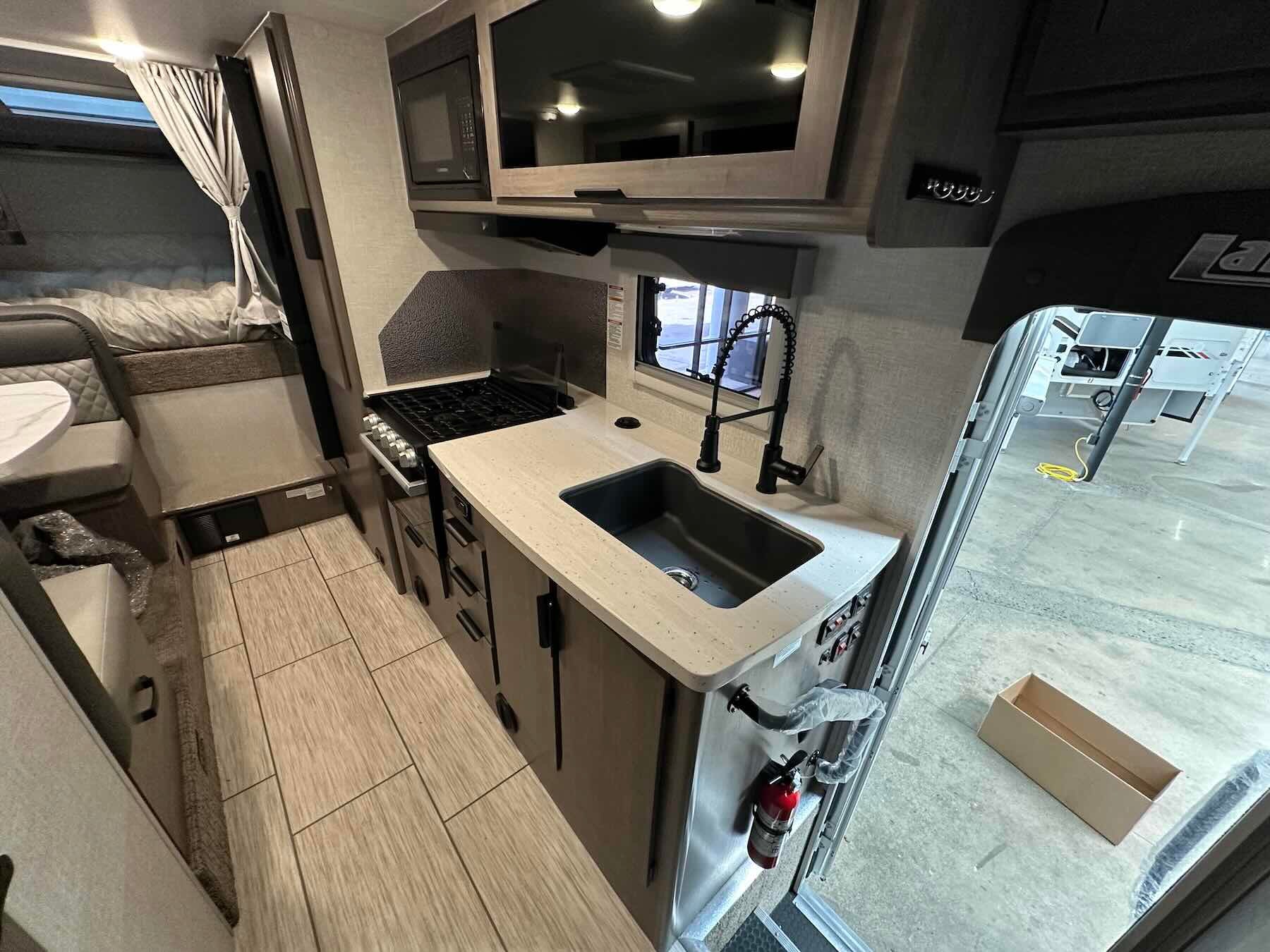 Interior of Lance 1191 showing kitchen with sink, stove, oven, microwave and cabinets, and cabover bed area in background.