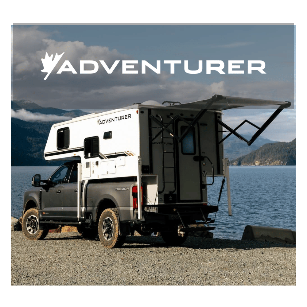 Truck with a camper shell labeled 'Adventurer' parked by a body of water with mountains in the background.