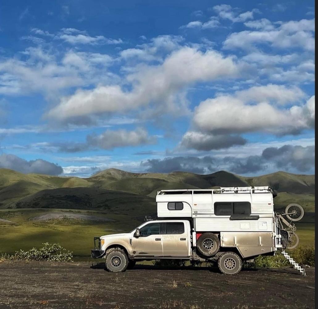 Dirty white truck with a white Alaskan flat bed cabover truck camper with roofracks stairs and bicycle on a dirt road with green hills and blue sky.