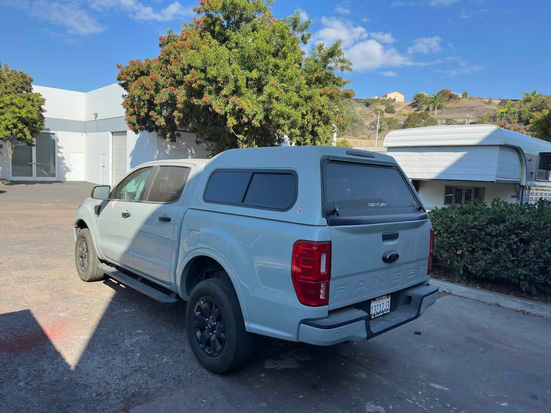 Light grey Ford Ranger with topper, rear driver corner, trees and building in background