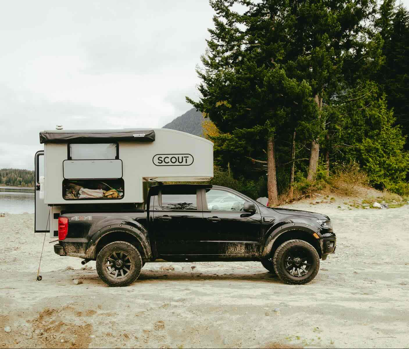 Black truck with a Scout camper shell parked on a sandy beach with trees and water in the background.