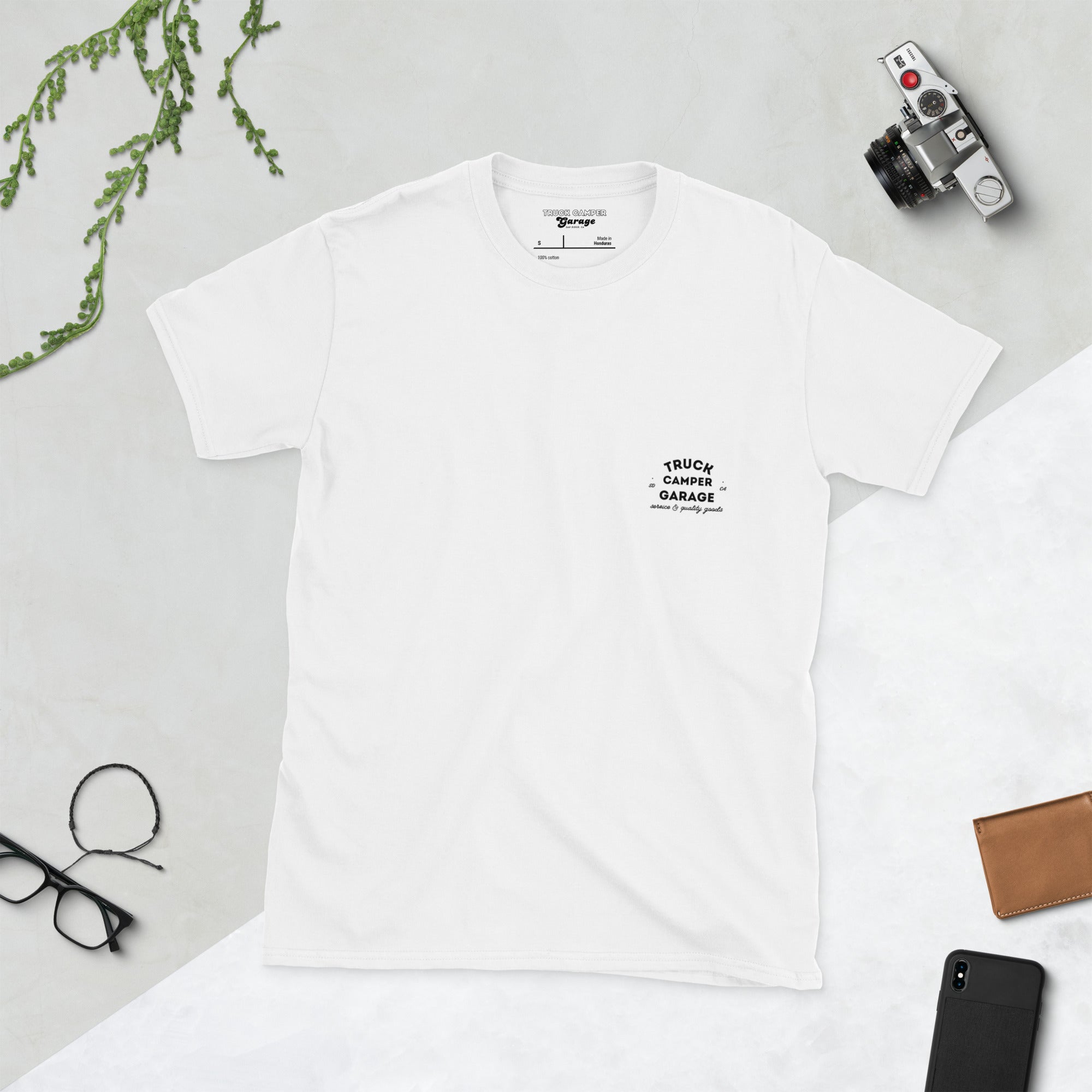 Front of white t-shirt with small truck camper garage logo on left chest plus camera glasses and phone in background